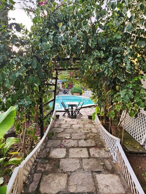 A stone pathway leading to a pool area with a table and chairs, surrounded by lush greenery and a white fence. - Flat em Maracaípe  (Ipojuca)