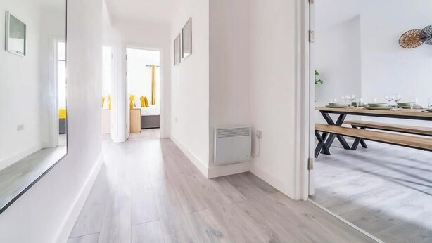 A modern hotel corridor with wooden flooring, a dining area with a wooden table and benches, and a bed with yellow bedding in the background. - Central Apartment | Contractors & Families | WiFi (Aldershot)