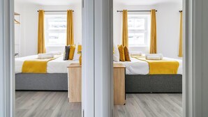 A modern bedroom with two beds, a wooden desk, and a window with yellow curtains. - Central Apartment | Contractors & Families | WiFi (Aldershot)