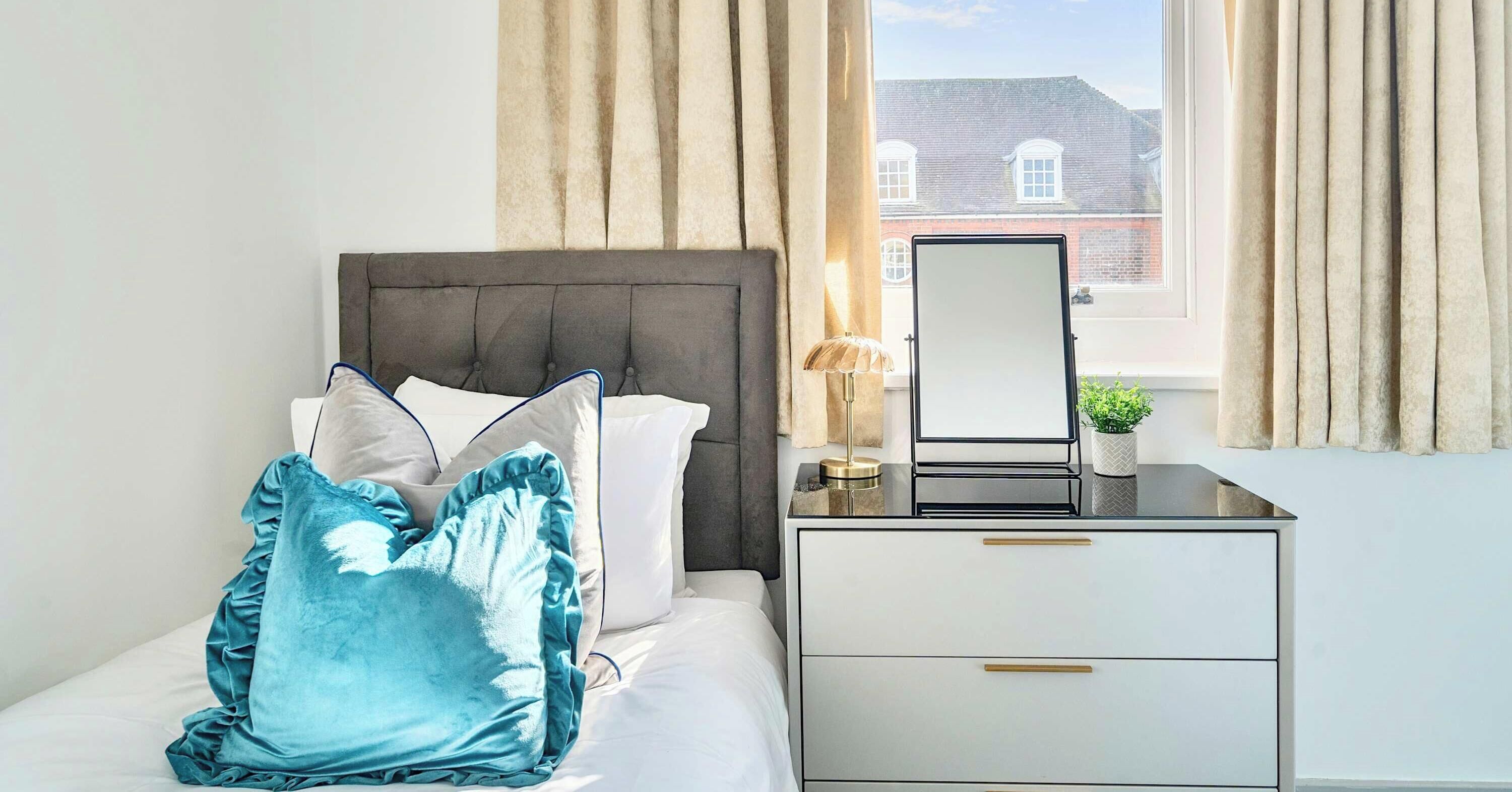 A neatly made bed with a grey headboard, a bedside table with a mirror, and a small potted plant.
