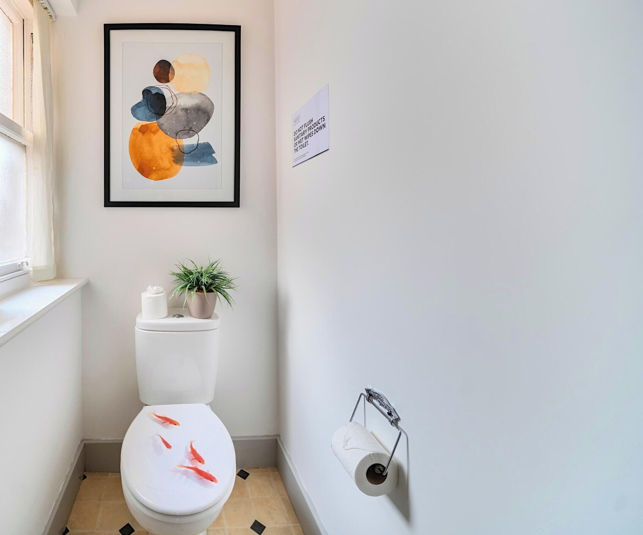 Un baño pequeño con inodoro, un cuadro abstracto enmarco, una planta en maceta y un dispensador de papel higiénico.