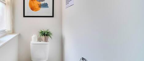 Un baño pequeño con inodoro, un cuadro abstracto enmarco, una planta en maceta y un dispensador de papel higiénico.