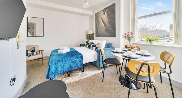 A modern hotel room with a bed, a sofa, a dining table with chairs, a large window, and a painting on the wall.