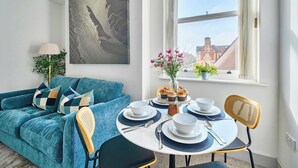 A modern living room with a teal sofa, a round dining table set for two, and a window with a view of buildings. - 1 Bed 1 Bath Apt, Contractors & Families, Parking (North Camp)
