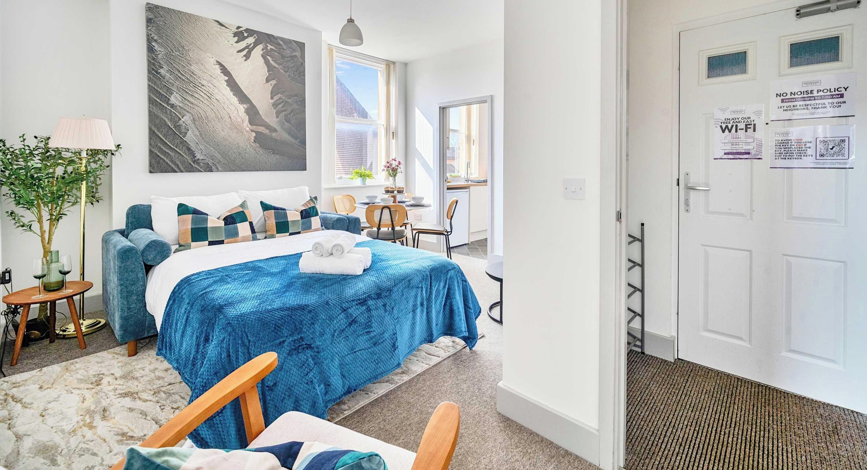 A modern bedroom with a large bed, a view of the city, and a door with a 'No Noise Policy' sign.