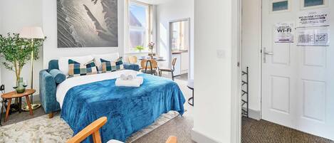 A modern bedroom with a large bed, a view of the city, and a door with a 'No Noise Policy' sign.