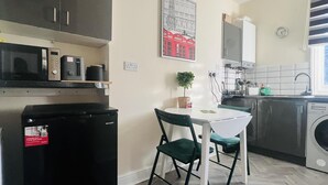 Une cuisine compacte équipée d’un four à micro-ondes, d’un petit réfrigérateur, d’un évier et d’un lave-linge. On y trouve une table avec une chaise et une plante en pot.