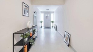 Un couloir avec une console noire, des plantes en pot, des tableaux encadrés et une porte blanche au fond.