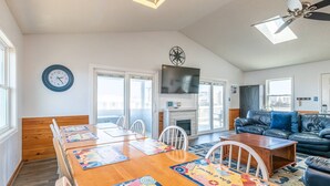 A spacious living room with a wooden dining table, white chairs, a black leather sofa, and a fireplace. - Southern Comfort - KDH MP 7.5, Semi-Oceanfront, 140 Yds to Beach, Hot Tub, Pets OK, Flex Stays (Kill Devil Hills)