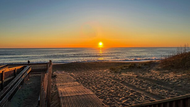 A beach with a wooden boardwalk leading to the water, a sunset in the background, and waves gently crashing onto the shore. - Southern Comfort - KDH MP 7.5, Semi-Oceanfront, 140 Yds to Beach, Hot Tub, Pets OK, Flex Stays (Kill Devil Hills)