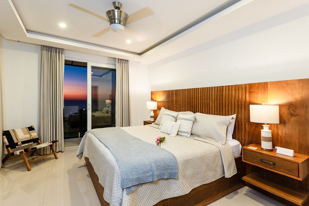 A modern hotel bedroom with a large bed, wooden headboard, bedside lamps, a nightstand, and a view of the city at sunset through the sliding glass door.