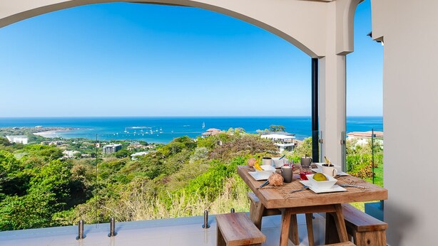 Un espace repas avec une table et des bancs, donnant sur un paysage côtier avec des bâtiments et de la végétation.