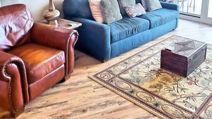 Un salon avec un canapé bleu, un fauteuil en cuir marron et un tapis à motifs. On y trouve trois tableaux accrochés au mur et un ventilateur de plafond.