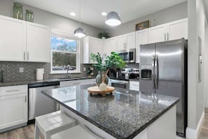 Cocina moderna con armarios blancos, encimeras de granito, electrodomésticos de acero inoxidable e iluminación colgante.