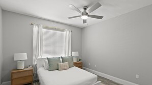 Une chambre à coucher comprenant un lit, deux tables de chevet, un ventilateur de plafond, une fenêtre avec des stores et un tapis.