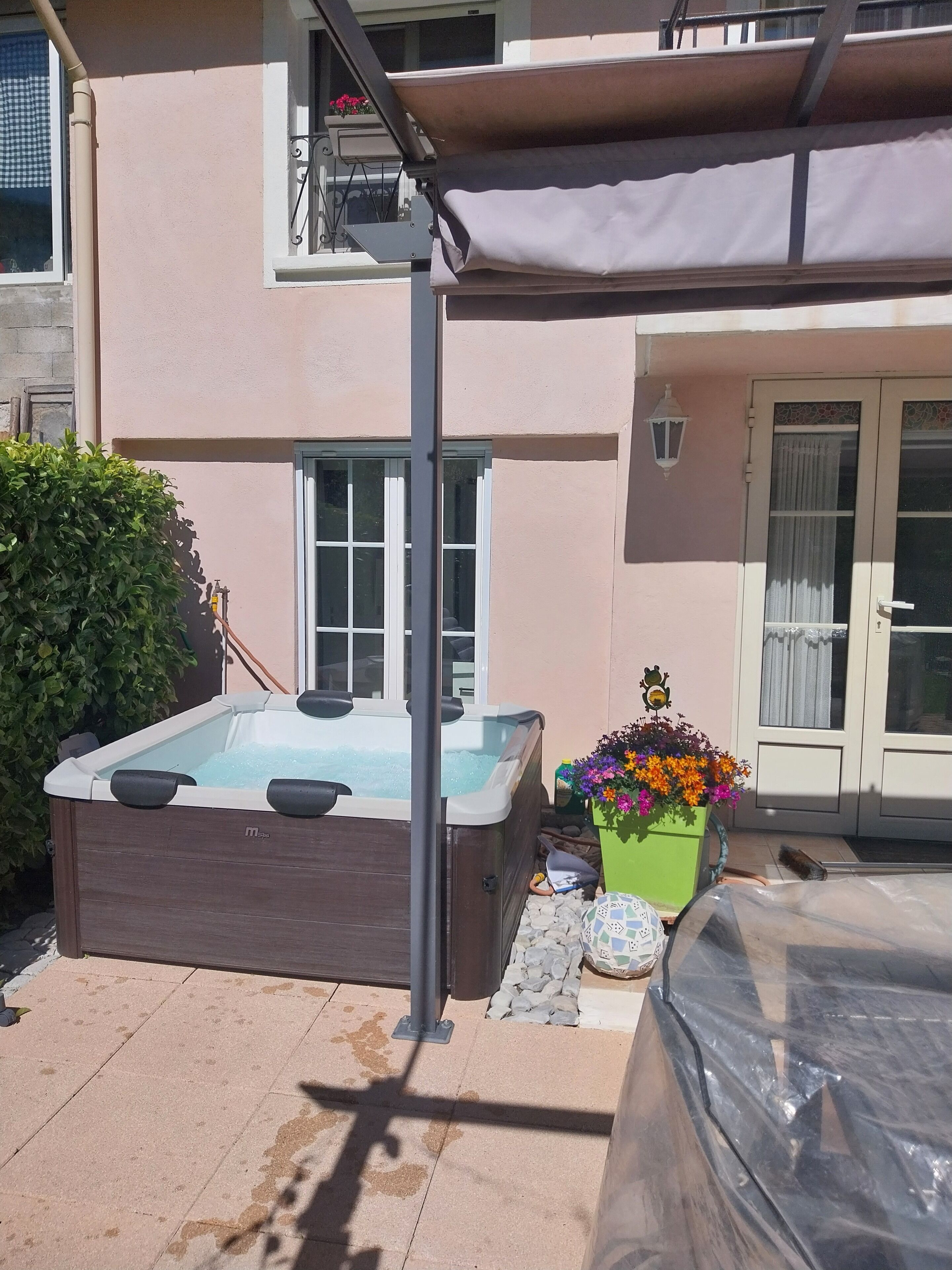 A hot tub in a backyard with a covered patio, a potted plant with colorful flowers, and a car covered with a tarp.