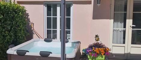 A hot tub in a backyard with a covered patio, a potted plant with colorful flowers, and a car covered with a tarp.