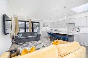 A modern kitchen with white cabinets, a central island, and bar stools. There is a grey sofa with patterned cushions in the living area, and a yellow sofa with yellow cushions.