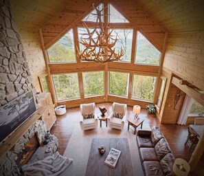 A spacious room with a high ceiling, large windows, a stone wall, and a fireplace. - Outstanding Views! 5BR/4.5 BA/ Sleeps 14, Hot-Tub, Pool Table! (Maggie Valley)