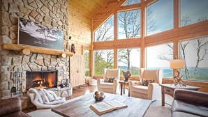 A cozy room with a stone fireplace, wooden walls, and large windows offering a view of the outdoors. - Outstanding Views! 5BR/4.5 BA/ Sleeps 14, Hot-Tub, Pool Table! (Maggie Valley)