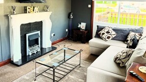 A living room with a fireplace, a television mounted on the wall, a sofa, and a window with a view of a fence and cars. - Pet Friendly - 6 Guests - Haven Lakeland (Grange-over-Sands)