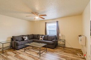 A living room with a sectional sofa, a glass coffee table, a ceiling fan, a window with curtains, and a wall-mounted air conditioning unit.