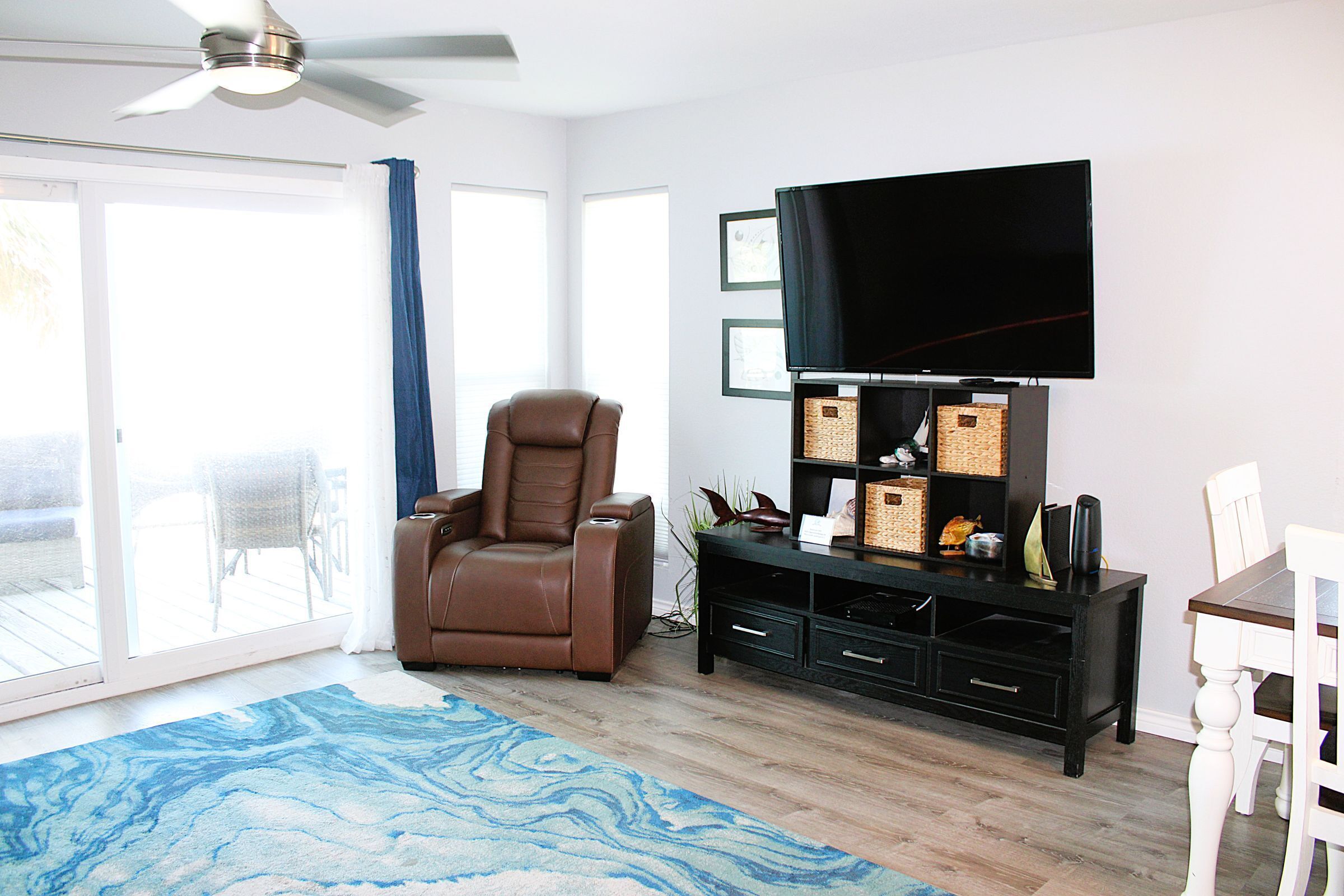 A living room with a ceiling fan, a brown leather recliner, a flat-screen TV on a black stand, a white dining table with chairs, and a blue patterned rug.