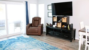 Ein Wohnzimmer mit Deckenventilator, einem braunen Ledersessel, einem Flachbildfernseher auf einem schwarzen Standfuß, einem weißen Esstisch mit Stühlen und einem blauen Teppich mit Muster.