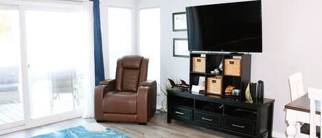 A living room with a ceiling fan, a brown leather recliner, a flat-screen TV on a black stand, a white dining table with chairs, and a blue patterned rug.