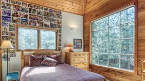 Une chambre avec une grande fenêtre, des murs en bois, un lit recouvert d’une courtepointe violette, une commode et un mur couvert de photos de famille.