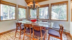 Un coin repas avec une table en verre, des chaises en bois, un lustre et de grandes fenêtres donnant sur la forêt.