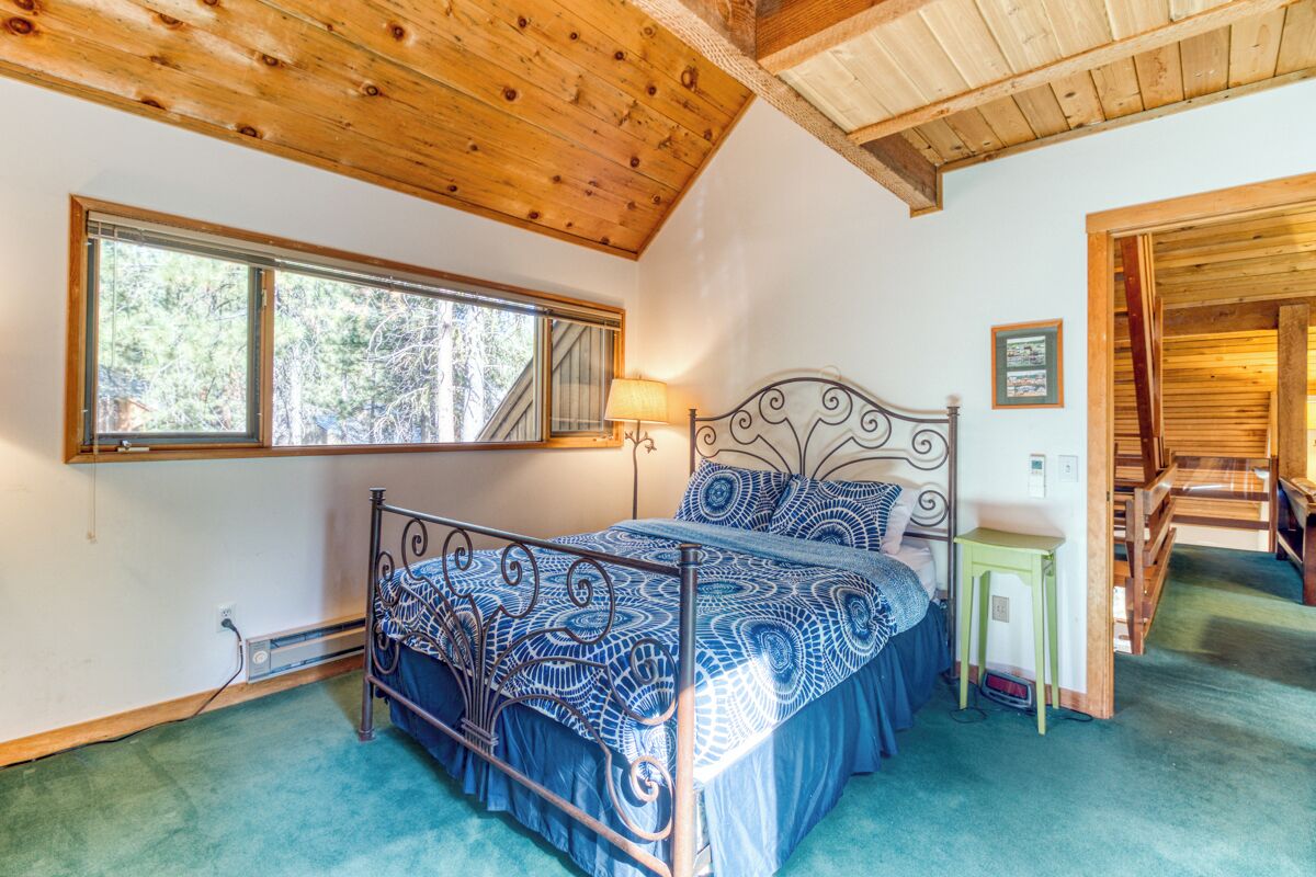 A bedroom with a metal bed frame, patterned bedding, a window with a view of trees, and a wooden ceiling.