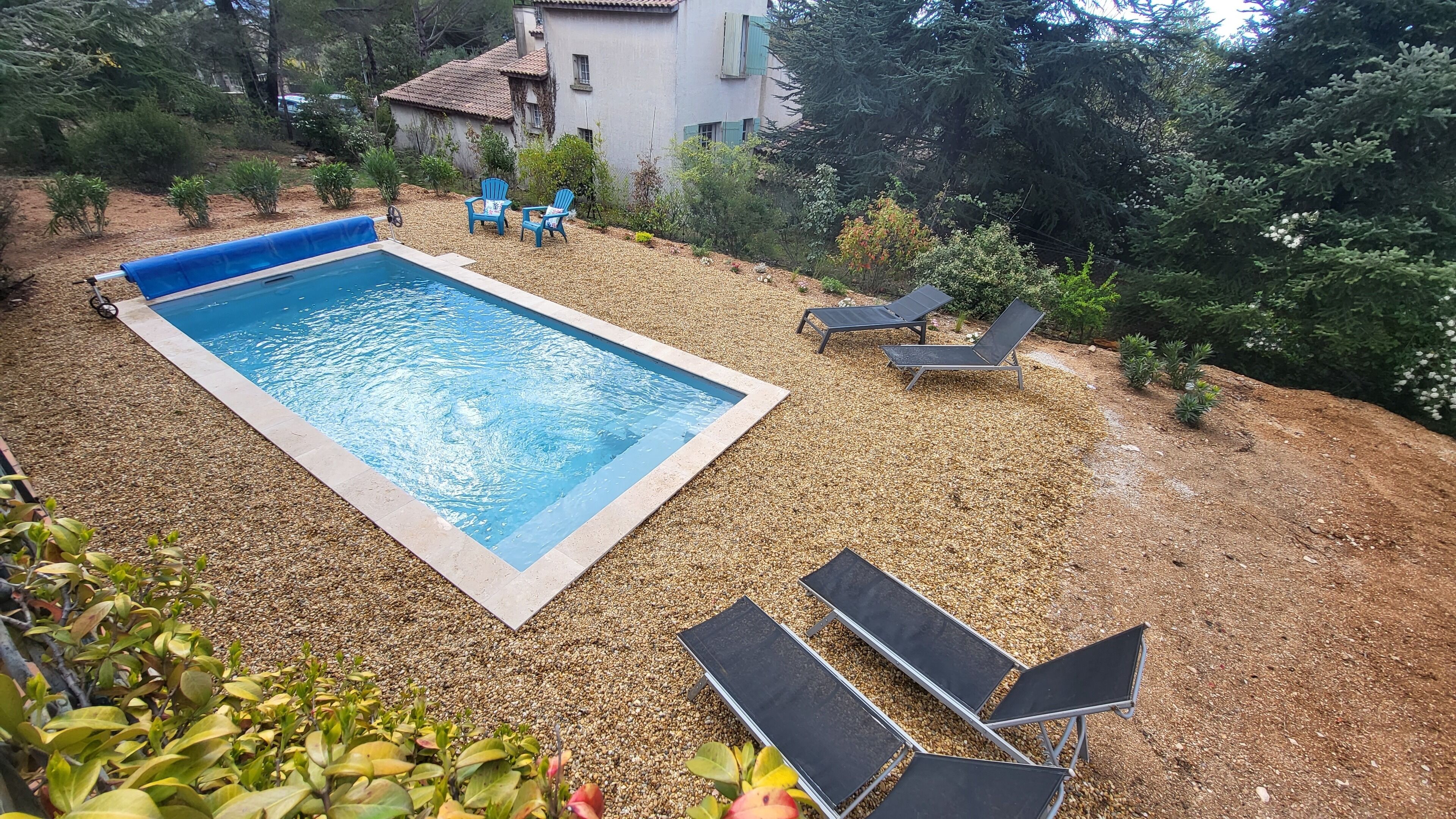 Ein rechteckiger Pool mit einer gefliesten Kante, umgeben von Kies. Zwei blaue Stühle und zwei Liegestühle mit schwarzen Kissen.