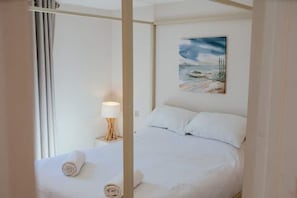 Een net opgemaakt bed met witte lakens en kussens, een bedlampje en een landschap schilderij aan de muur.