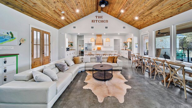 A spacious kitchen with a high wooden ceiling, a large dining area with wooden benches, and a modern white sofa set. - Pickle Houz, Pickleball Court, Pool, Golf@ Frio River Vacation Rentals (Concan)
