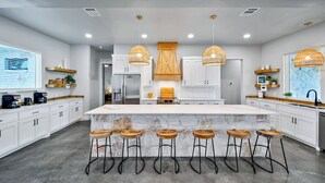 A modern kitchen with white cabinets, a large island with bar stools, pendant lights, and a stone backsplash. - Pickle Houz, Pickleball Court, Pool, Golf@ Frio River Vacation Rentals (Concan)