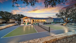 An outdoor tennis court with a building in the background. - Pickle Houz, Pickleball Court, Pool, Golf@ Frio River Vacation Rentals (Concan)
