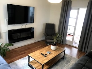 Een woonkamer met een flatscreen televisie aan de muur, een houten salontafel met een plant, een grijze fauteuil en een bank.