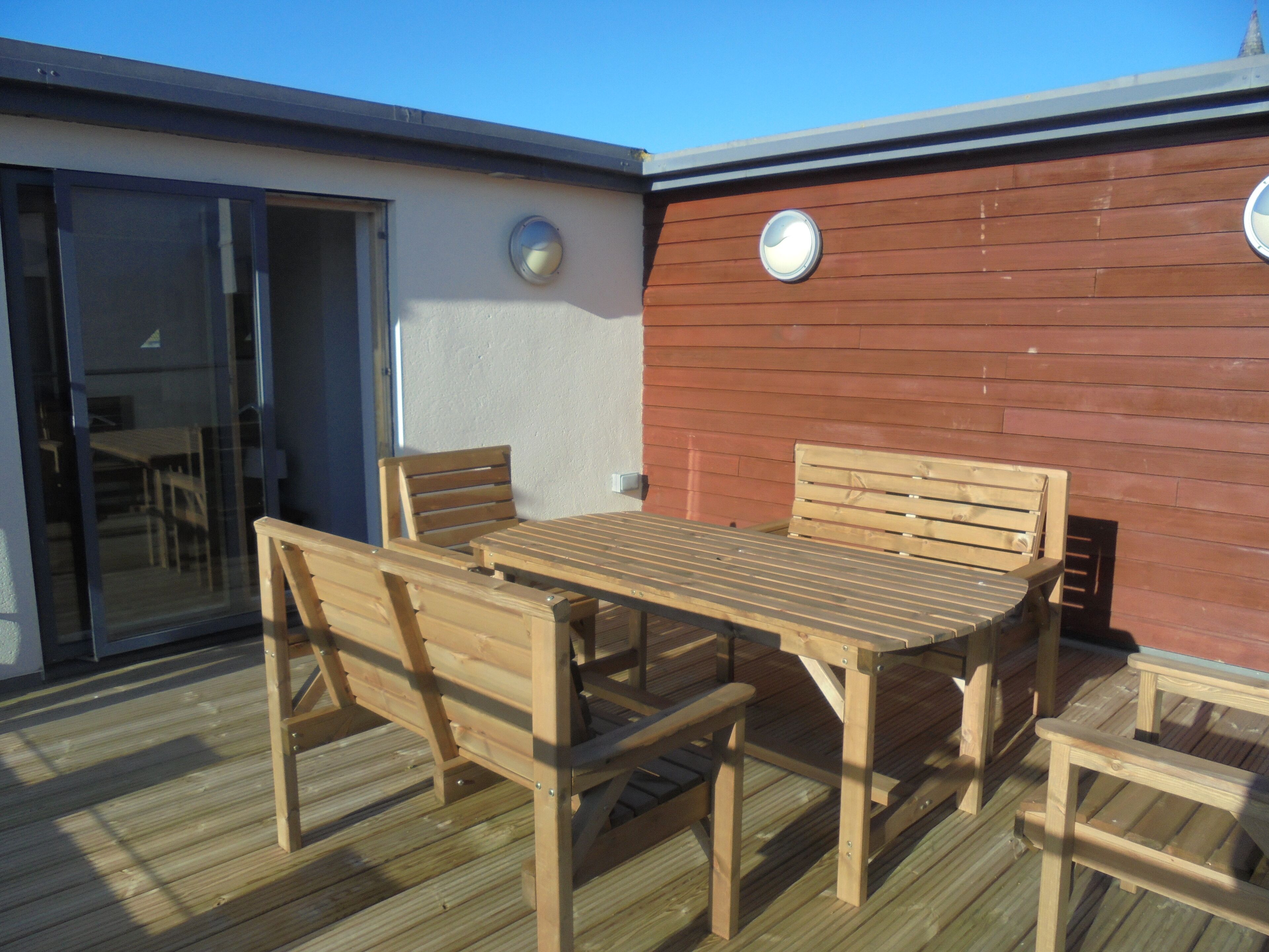 An outdoor wooden deck with a table and chairs, a glass door, and a red wall with two round windows.