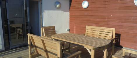 An outdoor wooden deck with a table and chairs, a glass door, and a red wall with two round windows.