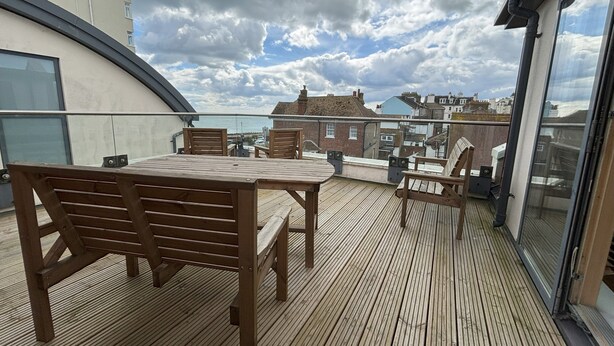Eine Holzterrasse mit Tisch und Stühlen, die auf einen Stadtblick mit Gebäuden und bewölktem Himmel blickt.