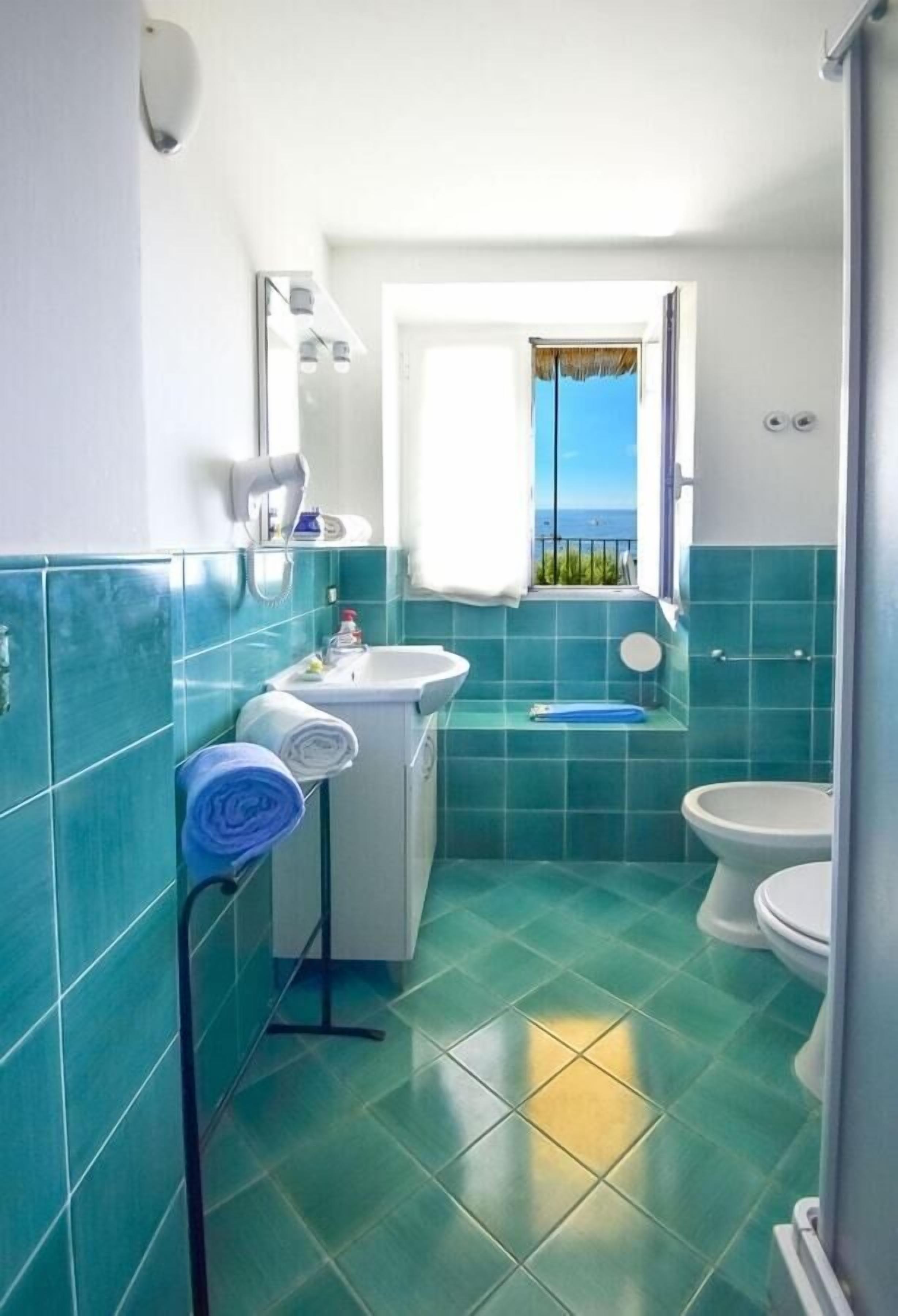 Et lille badeværelse med vask, toilet og bruser. Der er teal farvede fliser og et vindue med udsigt.