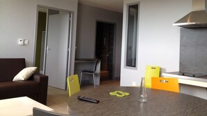 A modern living room with a sofa, dining table, chairs, and a kitchen area with a stove. - Résidence Apparten Ville Appartement 6 Personnes (Saint-Flour)