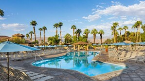 A pool area with lounge chairs and umbrellas, surrounded by palm trees and a clear sky.