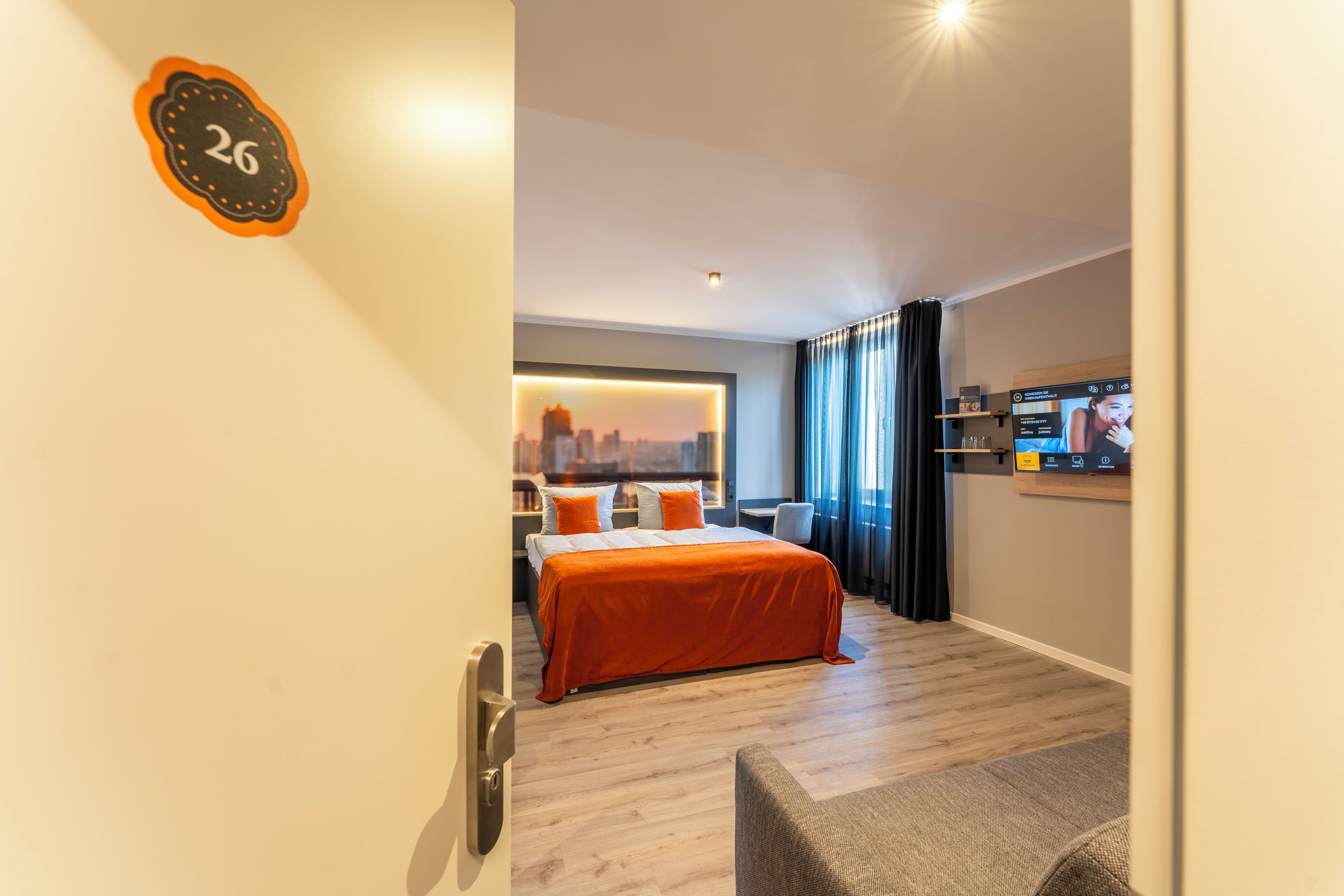 A hotel room with a bed, a cityscape view, a TV, and a door with a number.