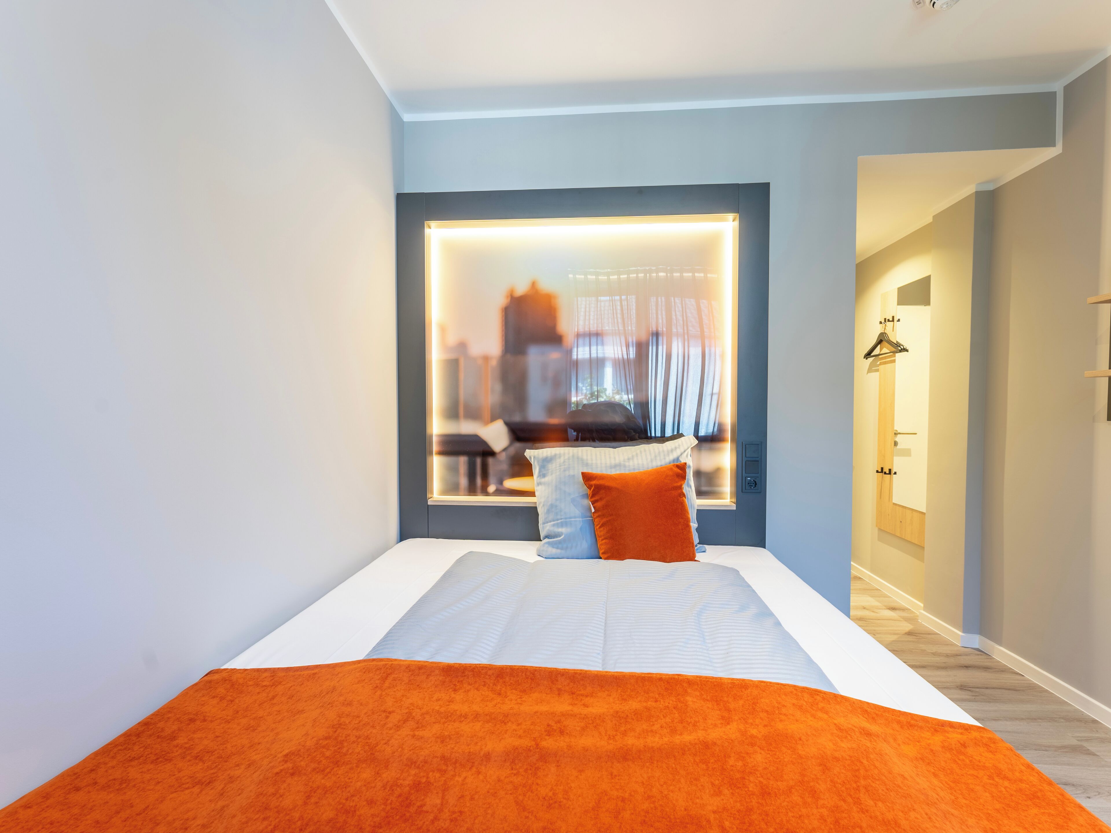 A modern hotel room with a large bed, a view of the city through a window, and a hallway leading to the bathroom.