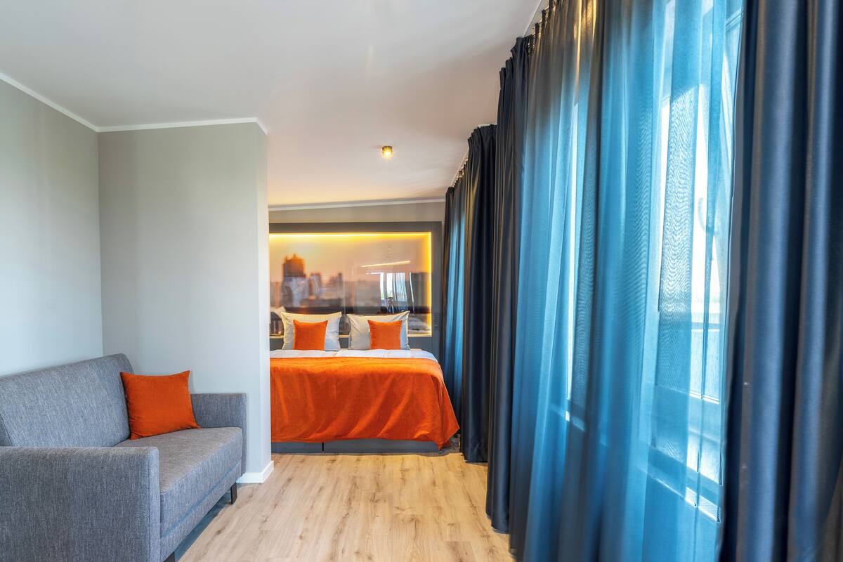 A modern hotel room with a grey sofa, an orange bedspread, and a city view through large windows with blue curtains.