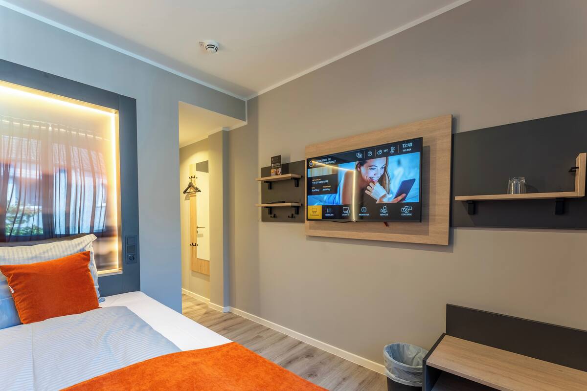 A modern hotel room with a bed, a TV mounted on the wall, a wooden bench, and a trash bin.