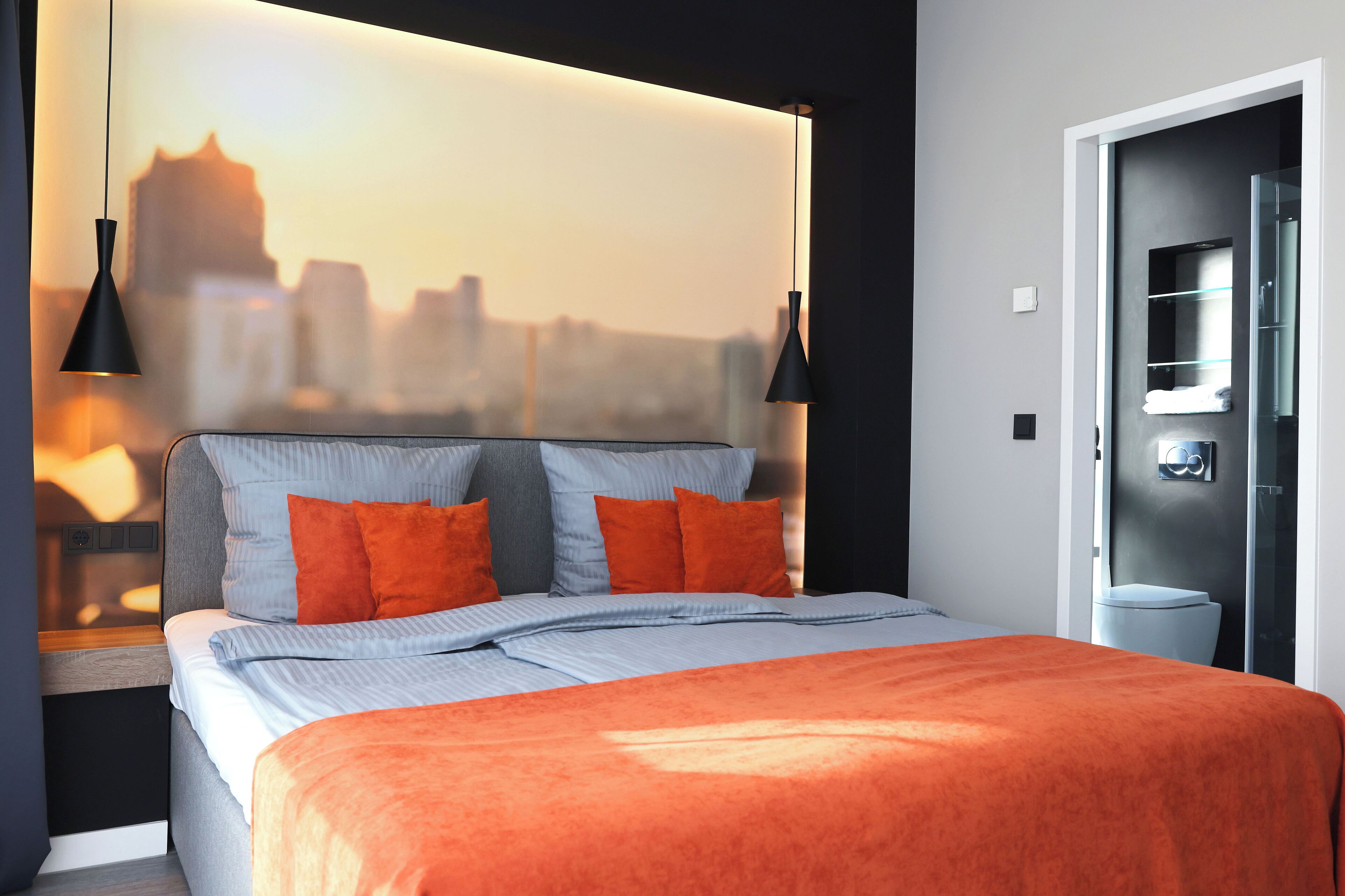 A modern bedroom with a cityscape view, a bed with orange bedding, and two pendant lights.