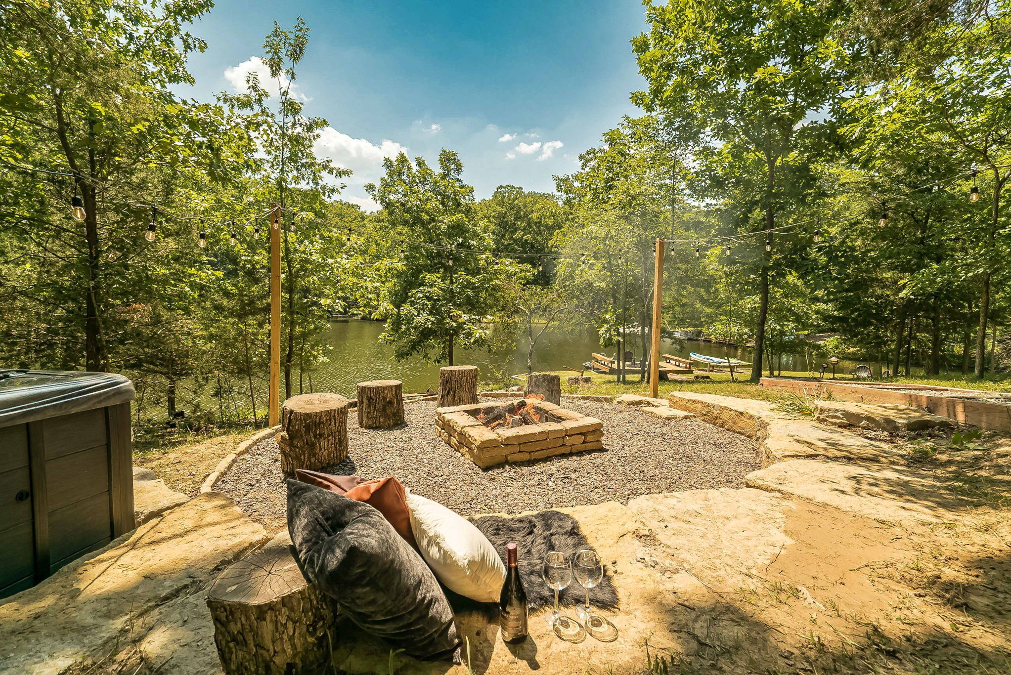 A hot tub, fire pit, and outdoor seating area surrounded by trees and a lake.